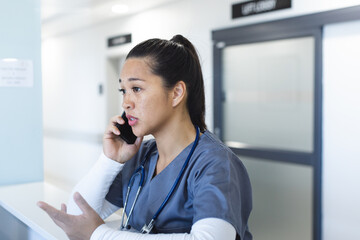 Biracial female doctor talking on smartphone at reception desk in hospital