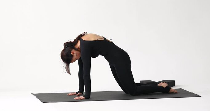 A yoga instructor performs a variation of the Marjariasana exercise, a cat pose with an up and down bend, training in a black long-sleeve jumpsuit on a mat in a studio against a light background