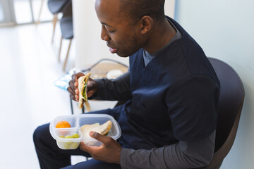 African american male doctor wearing scrubs eating takeaway food in waiting room in hospital