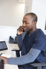 African american male doctor talking on smartphone and taking notes at reception desk in hospital