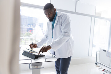 African american male doctor using tablet and taking notes in hospital room, copy space