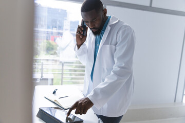 African american male doctor using tablet and talking on smartphone in hospital room