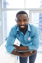 Portrait of happy african american male doctor holding tablet at reception desk in hospital