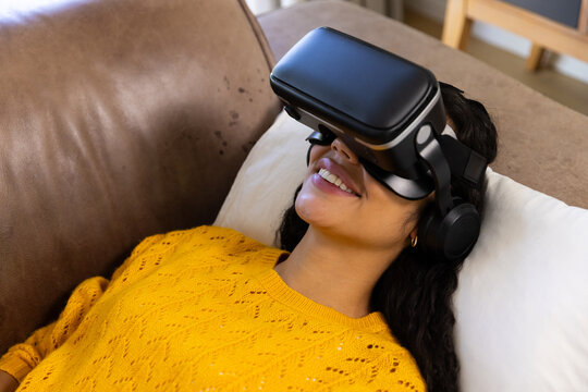 Happy biracial woman lying on sofa and using vr headset at home