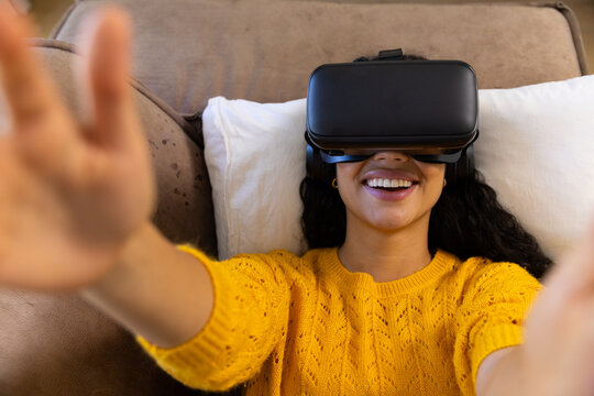Happy biracial woman lying on sofa and using vr headset at home