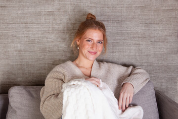 Chica rubia pelirroja sonriendo en la comodidad de su casa sentada en el sofá del salón con una manta entre sus piernas