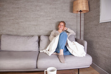Joven mujer sonriente disfrutando de la comodidad de su hogar, relajada en un sofá gris con manta...