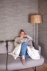 Joven mujer sonriente disfrutando de la comodidad de su hogar, relajada en un sofá gris con manta...