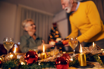 Close up of christmas ornaments and candles on christmas table on christmas eve with senior couple in a blurry background.