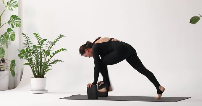 Woman standing in a plank and leaning her hands on small sports bricks, performs an exercise to strengthen the muscles of the anterior thigh, extends and bends her leg, trains on the mat in the studio