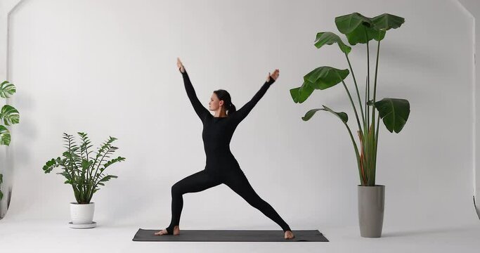 Woman performing Virabhadrasana exercise, warrior pose number one and two, training in black sports overalls while standing on a mat in a room with flowers