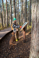 Tourist woman walking with dog on ecotrope reading information about plants on stand in Scandinavian nature park forest. Pet owner traveling enjoying woodland with pine trees. Travel tourism concept.