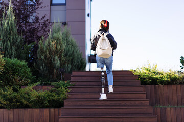 Young woman student with prosthetic leg walking upstairs in city. Disabled woman with prosthetic...