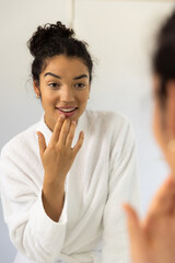 Happy biracial woman in bathrobe looking in mirror in sunny bathroom
