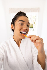 Portrait of biracial woman in bathrobe brushing teeth in sunny bathroom