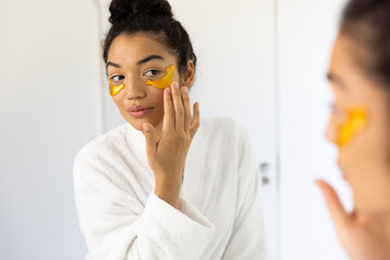 Biracial woman in bathrobe applying eye mask in sunny bathroom