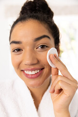 Portrait of happy biracial woman in bathrobe cleansing her face in sunny bathroom