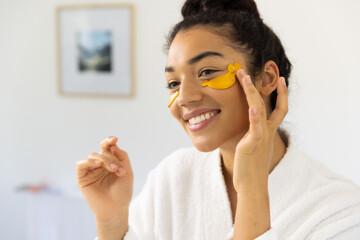 Happy biracial woman in bathrobe applying eye mask in sunny bathroom