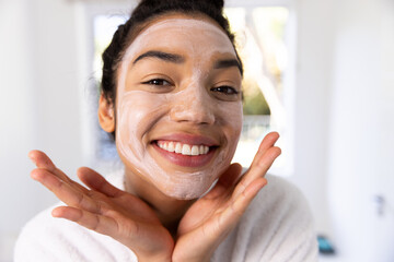 Portrait of happy biracial woman in bathrobe with face mask in sunny bathroom