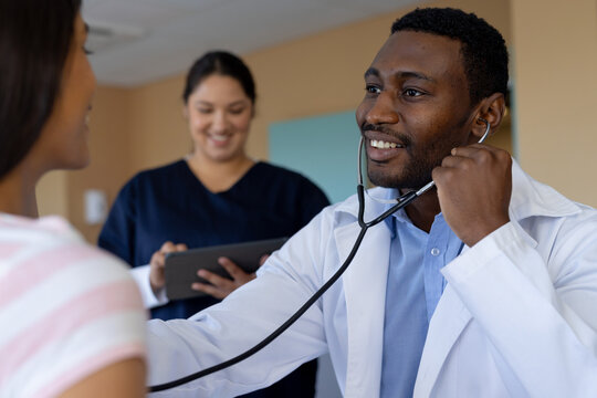 Happy diverse male doctor testing female patient using stethoscope in hospital room