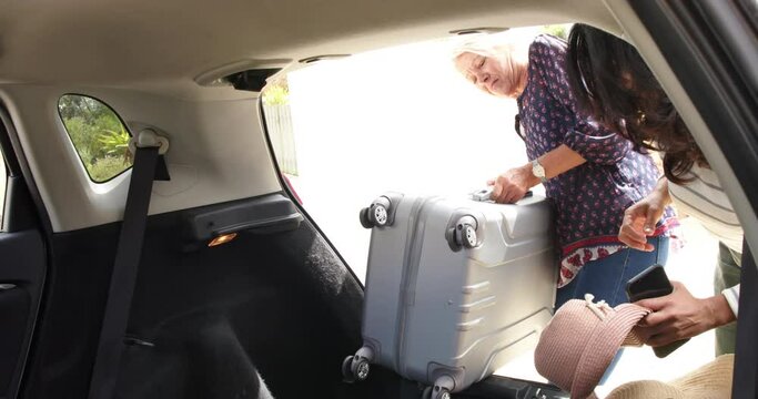 Two happy diverse senior women putting suitcase into car on sunny day, slow motion