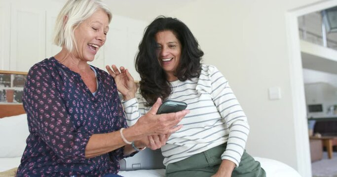 Two happy diverse senior women using smartphone and laughing in sunny bedroom, slow motion