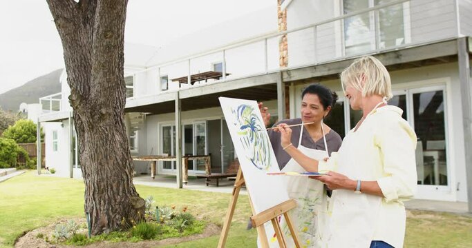 Two happy diverse senior women painting picture and laughing in sunny garden, slow motion