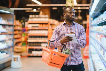 african american man standing in supermarket choosing products while reading notes at notebook