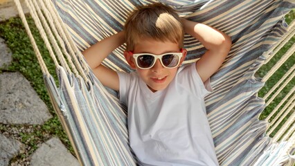 At peace in a garden a boy in sunglasses swings and lounges in a hammock, expressing the sensations of summertime, the innocence of childhood, calmness, and the magic of summer holidays