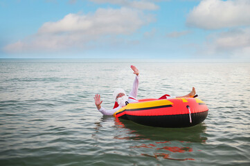 Santa in red suspenders floats on an inflatable boat against the background of the sea.