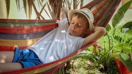 A boy sways and rests in a hammock at garden, portraying the sensations of summertime, treasured childhood memories, and the relaxation of a vacation