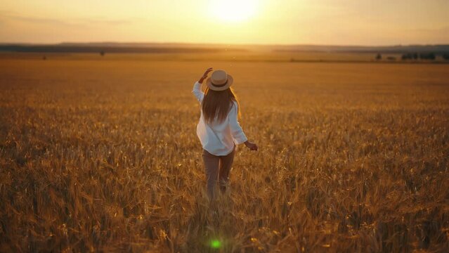 Carefree woman in straw hat running on wheat field feeling freedom, harmony, lightness and serenity, back view. Smiling blonde female enjoying nature resting on farm, outdoors activity. Sunset time.