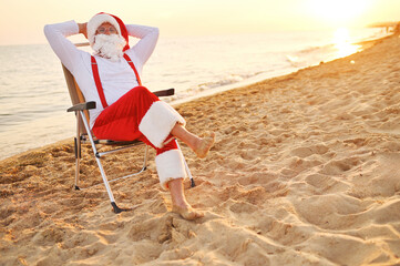 Santa barefoot in red suspenders sitting on a chaise lounge resting against the background of the...