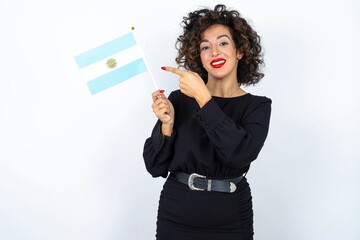 Young beautiful woman with curly hair wearing black dress and holding an Argentina flag and pointing at it. 