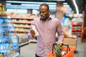 Surprised African-American man looks at receipt total in sales check holding paper bag - obrazy, fototapety, plakaty