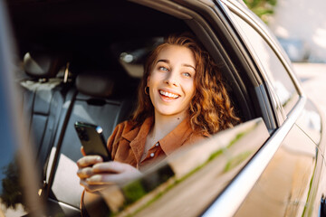 Portrait of a happy taxi passenger with a phone in her hands. A young woman with a smartphone chats in the back seat of a car. Transport concept. Lifestyle.