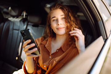 Portrait of a happy taxi passenger with a phone in her hands. A young woman with a smartphone chats in the back seat of a car. Transport concept. Lifestyle.