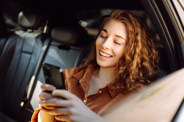 Portrait of a happy taxi passenger with a phone in her hands. A young woman with a smartphone chats in the back seat of a car. Transport concept. Lifestyle.