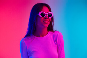 Beautiful young woman posing in sunglasses against gradient pink blue studio background in neon light. Positivity. Concept of human emotions, lifestyle, youth culture, facial expression