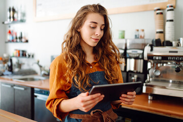 A young woman in an apron with a digital tablet at the bar counter of a coffee shop takes an order. Small business, technology concept.