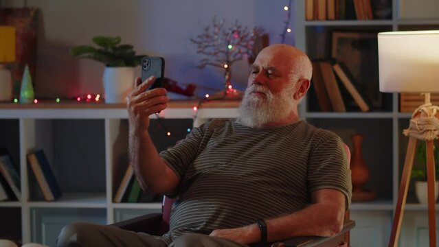 An elderly man in good health and positive mood sits in the room in an armchair, uses a smartphone and video call conversation. A human waves hand as greeting sign, smiles, talks animatedly and