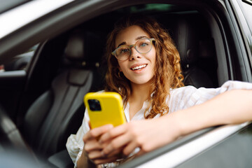Portrait of a beautiful curly woman driving a car with a phone in her hands. Happy woman uses...