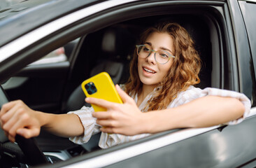 Fototapeta na wymiar Portrait of a beautiful curly woman driving a car with a phone in her hands. Happy woman uses mobile navigation for car movement. Concept of transport, mobility.