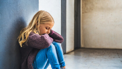 A young girl sits alone with a sad feeling at school near the wall. Offended child abandoned in the corridor and bent against a brick wall. Bullying, discrimination with copy space