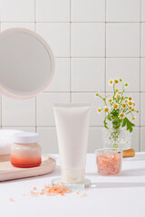 Unlabeled tube and jar decorated with a bowl of pink himalayan salt in the bathroom. A rounded mirror and flower pot featured. Branding mockup