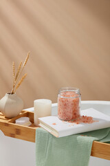 Wooden bathtub tray with a flower pot, foot brush, candle, book and a jar pink himalayan salt placed on. Pink himalayan salt blast away dead cells for softer, brighter-looking skin