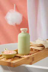 A bottle in pastel color placed on a bathtub tray with a dish containing pink himalayan salt and a towel with foot brush. White bath sponge hanging on the wall. Empty label for branding mockup