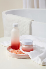 Rounded marble tray with a blank label bottle and jar placed on. A handful of pink himalayan salt displayed. Background with a bathtub with white towel hanging on. Branding mockup