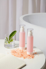 Pink bottles without label standing on the table with many pink himalayan salt, a notebook and a cup of green leaves. Skin care concept with empty label