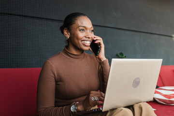 Cheerful young black woman talking on smartphone while sitting with laptop